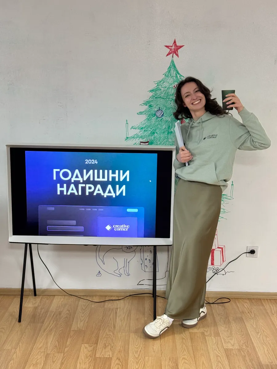 Woman smiling and holding a cup and papers next to a screen showing '2024 ГОДИШНИ НАГРАДИ' with a drawn Christmas tree on the wall.