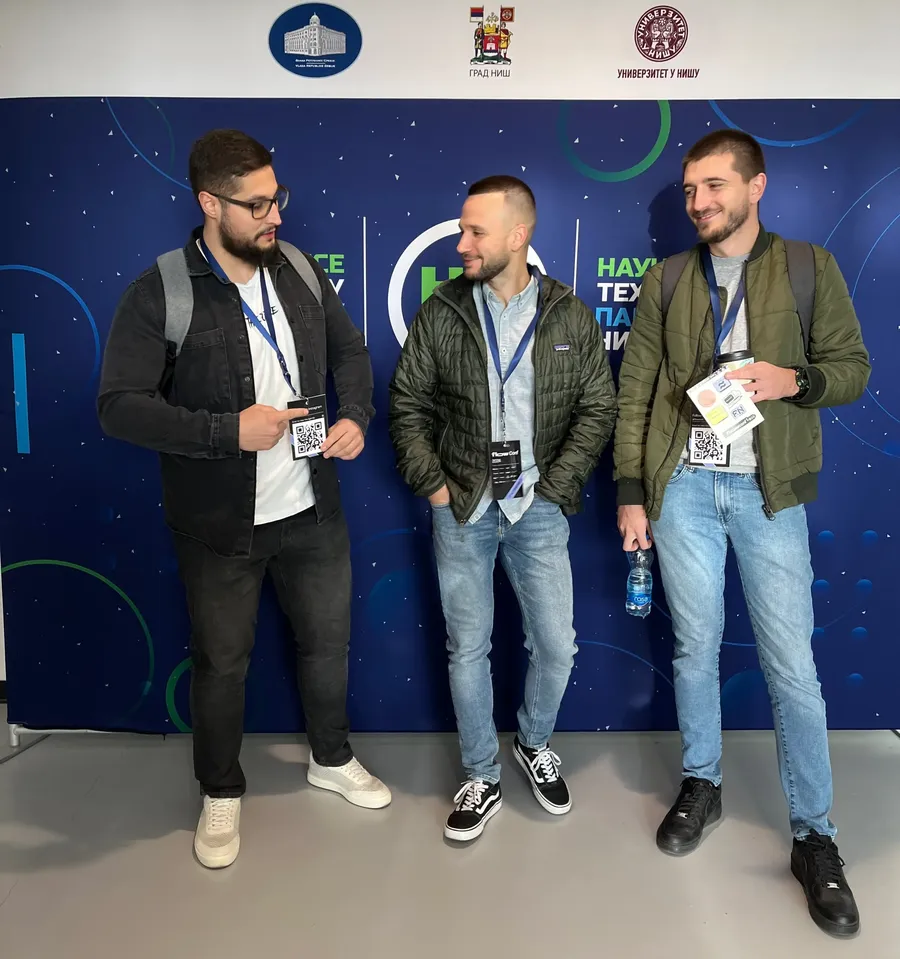 Three men with conference badges stand and chat in front of a blue backdrop with logos and text.