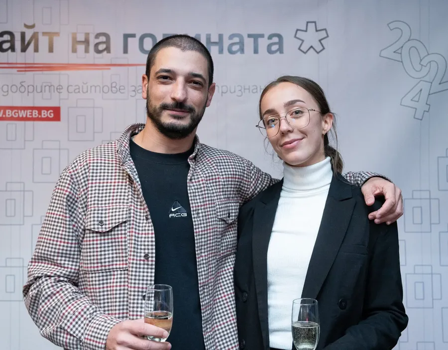 Man in checkered shirt and woman in black blazer holding champagne glasses, posing together at an event.