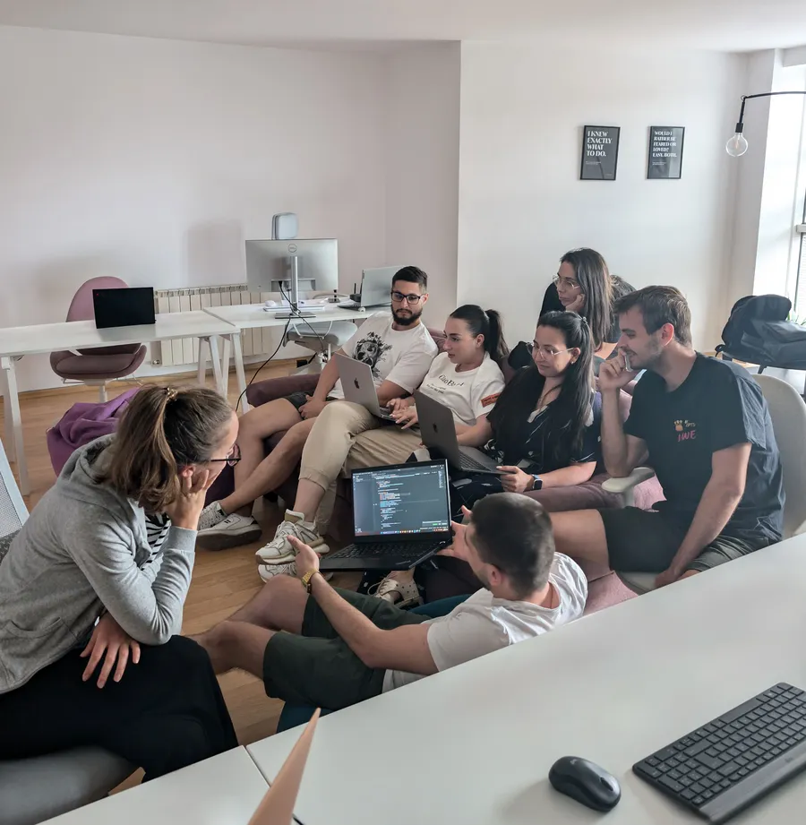 A group of seven young adults gathered in a casual office, focused on laptops with coding on screen during a collaborative meeting.